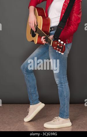 Seitenansicht der unteren Hälfte einer nicht erkennbaren Gitarristin, die sechs-saitige Akustikgitarre spielt. Der stilvolle junge Musiker trägt rotes Leder Stockfoto
