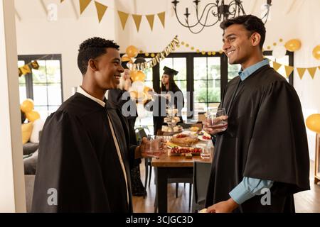 Absolventen in Mützen und Kleidern feiern mit Getränken zu Hause und lächeln fröhlich Stockfoto