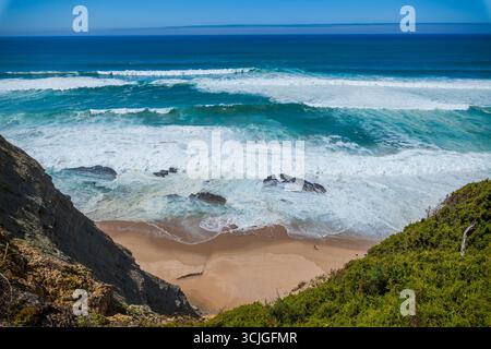 Ein atemberaubender Blick auf die Küste von oben, mit türkisfarbenen Wellen, die auf einen Sandstrand zwischen felsigen Klippen und grünen Sträuchern unter einem klaren Wasser stürzen Stockfoto