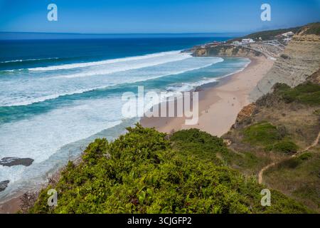 Ein weites Küstenpanorama mit einem Sandstrand mit stürzenden Wellen, zerklüfteten Klippen, gewundenen Wegen und üppigem Grün unter einem leuchtend blauen Himmel Stockfoto
