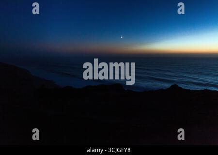 Eine ruhige Küstenszene in der Abenddämmerung mit einem Halbmond, der über dem ruhigen Ozean leuchtet, Silhouetten von Klippen und einem verlaufenden Himmel, der von tiefem Blau übergeht Stockfoto