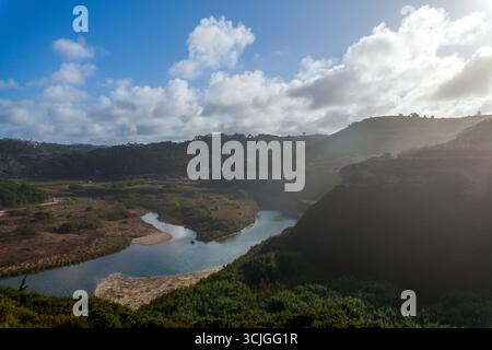 Ein sich windender Fluss fließt durch ein üppiges Tal, umgeben von sanften Hügeln, dichter Vegetation und dramatischen Klippen unter einem hellblauen Himmel mit verstreuten Flächen Stockfoto