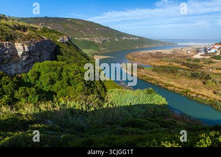 Ein ruhiger Fluss schlängelt sich durch ein üppiges grünes Tal, das von felsigen Klippen flankiert wird, und führt zu einem sandigen Ufer und dem Meer unter einem klaren blauen Himmel auf Praia da Fo Stockfoto