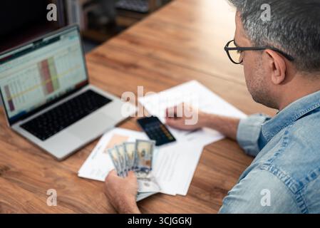 Grauhaariger Mann hält Dollarfan über Stromrechnungen am Laptop in den Haushaltsausgaben des Firmenbüros Stockfoto