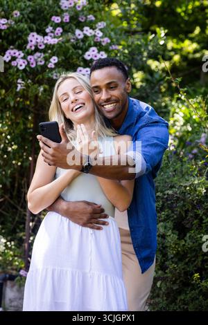 Paare feiern Verlobung, lächeln und machen Selfie im Garten mit dem Smartphone Stockfoto