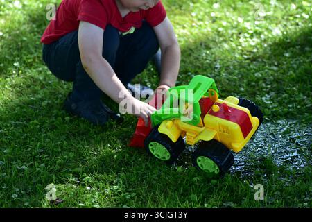 Kinder in der Hocke und spielen mit einem bunten Plastikspielzeugtraktor auf nassem Gras mit Wasser, Sommer-Outdoor-Spiel. Kindliche Vorstellungskraft und lustige Aktivität. Stockfoto