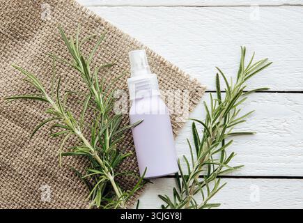Hausgemachtes Rosmarin Salvia rosmarinus Wasser aus Rosmarinblättern zur Haar- und Hautpflege. Eine lila Sprühflasche zwischen Rosmarinzweigen auf weiß Stockfoto