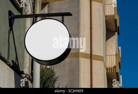 Leere runde Ladenschilder auf einer Metallhalterung, die vor einem Gebäude mit moderner beigefarbener Fassade hängt. Der klare blaue Himmel im Hintergrund Stockfoto