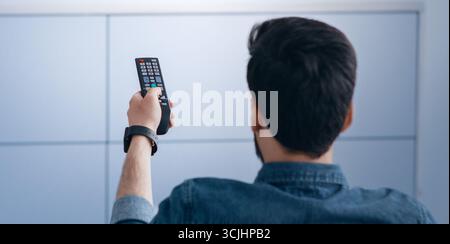 Mann, der Fernsehen guckend, den Plasma-Flachbildschirm-Fernseher anschminkend, die Fernbedienung auf den leeren Fernsehbildschirm an der weißen Wand gerichtet Stockfoto