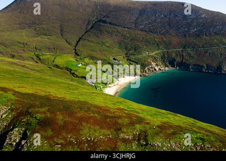 Aus der Vogelperspektive auf Keem Beach mit ruhigem blauem Atlantikwasser, Autos, die entlang der Straße geparkt werden, und einer gewundenen Bergstraße im County Mayo Stockfoto
