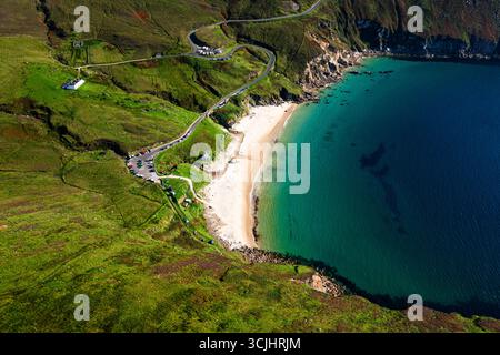 Aus der Vogelperspektive auf Keem Beach mit ruhigem blauem Atlantikwasser, Autos, die entlang der Straße geparkt werden, und einer gewundenen Bergstraße im County Mayo Stockfoto