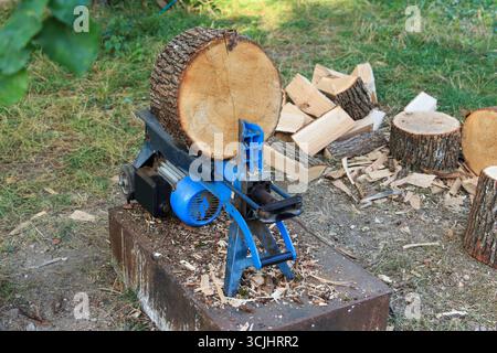 Ein blauer elektrischer Holzspalter befindet sich auf einer Holzplattform. In der Nähe sind Baumstämme gestapelt, einige davon sind bereits in kleinere Stücke aufgeteilt. Szene ist gesetzt o Stockfoto