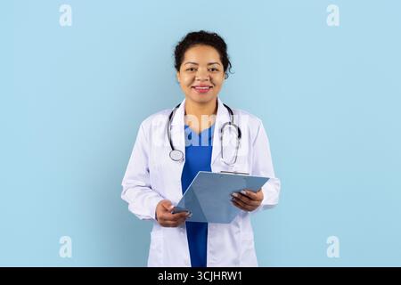 Schwarze Ärztin im weißen Mantel mit Stethoskop, das Clipboard im Atelier hält Stockfoto