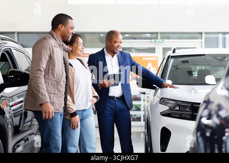 Ein lächelndes schwarzes Paar mit einem Autohändler, der den neuen SUV in einem modernen Showroom untersucht Stockfoto