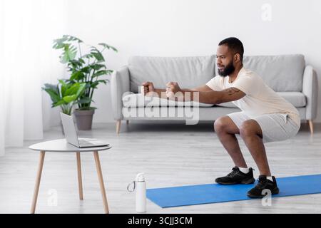 Konzentrierter junger schwarz bärtiger Athlet Mann in weißen Sportkleidung hockt im Wohnzimmer Innenraum, Profil Stockfoto