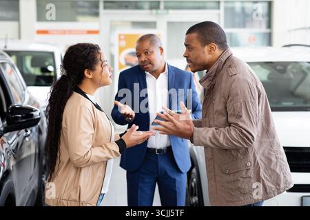 Ein junges afroamerikanisches Paar streitet im Autohaus, während der Händler zusieht Stockfoto