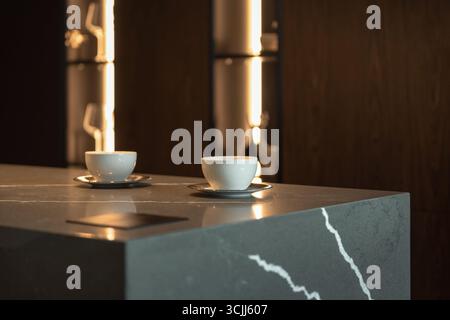 Zwei Tassen Kaffee auf einem Tisch. Zwei kleine weiße Tassen auf einem grauen Steintisch, warme Beleuchtung im Hintergrund. Stockfoto