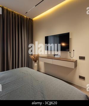 Wandmontierter Fernseher im Schlafzimmer. Großer Flachbildschirm an einer cremebeigen Wand, Zimmer mit beleuchteter Decke und braunem Vorhang. Stockfoto