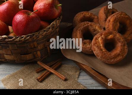 Apfelwein-Donuts. Frisch gebackene Donuts und ein Korb mit Gala-Äpfeln mit Zimtstangen auf einem rustikalen weißen Küchentisch. Stockfoto