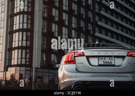 Rückansicht des silbernen Autos gegen Wohngebäude - geringer Kamerawinkel. Rückansicht der Volvo S60 Limousine in einer Stadt bei Sonnenuntergang, modernes Gebäude hinten Stockfoto