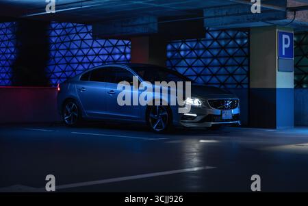 Volvo S60 in Parkgarage mit blauem Licht. Seitenansicht der schwedischen Limousine bei Nacht – Scheinwerfer eingeschaltet, silberne Karosserie reflektiert blaue Lichter. Stockfoto