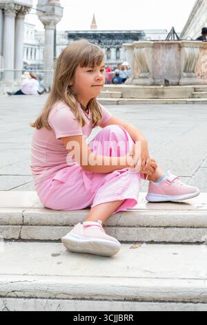 Ein 8-jähriges Mädchen in rosa Kleidung sitzt auf den Stufen eines venezianischen Platzes Stockfoto