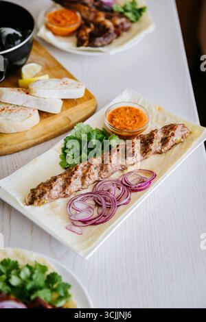 Traditioneller Kebab mit Kräutern, roten Zwiebeln und Sauce auf Lavash, serviert auf einem weißen Teller Stockfoto