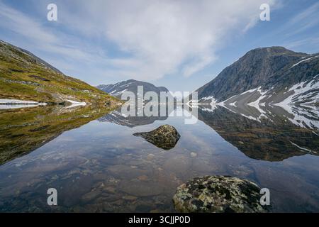 Norwegischer Bergsee mit kristallklarem Wasser, das schneebedeckte Gipfel und felsige Hänge unter hellem Himmel reflektiert Stockfoto