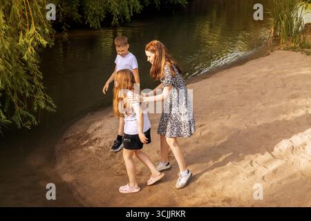 Drei kaukasische Kinder, zwei Mädchen und ein Junge, spielen an einem sonnigen Sommertag am Sandrand eines ruhigen Flusses. Für Themen des Kindes Stockfoto