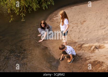 Drei kaukasische Kinder, zwei Mädchen und ein Junge, spielen an einem sonnigen Sommertag am Sandrand eines ruhigen Flusses. Für Themen des Kindes Stockfoto