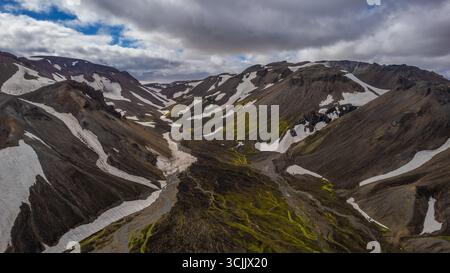 Blick aus der Vogelperspektive auf die vulkanischen Berge in Island mit schneebedeckten Gipfeln, einem lebendigen grünen Moostal und einem sich windenden Fluss, der durch die raue Landschaft führt Stockfoto