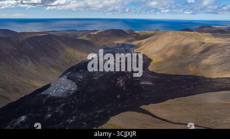 Blick aus der Vogelperspektive auf das vulkanische Tal Islands mit dunklem Lavastrom, der durch karge, braune Hügel schneidet, und Fernsicht auf die Atlantikküste Stockfoto