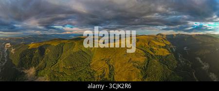 Sanfte grüne Hügel und bewaldete Berge in Europa unter stimmungsvollem Himmel mit dramatischen Wolken und Sonnenlichtansichten aus der Luft Stockfoto