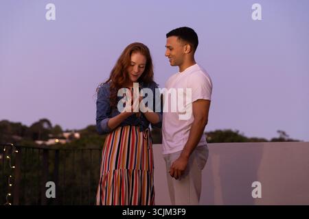Ein junges Paar genießt den Sonnenuntergang auf dem Dach, eine Frau zeigt etwas auf dem Smartphone Stockfoto