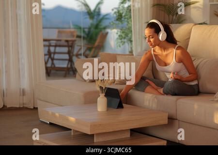 Yogi zu Hause, der geführte Yoga-Onlinekurs auf einem Tablet vorbereitet und bei Sonnenuntergang zu Hause auf einem Sofa sitzt Stockfoto