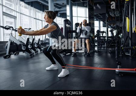 Reife Frau, die Squats mit einem Gummiband macht, mit einem sportlichen Mann im Hintergrund Stockfoto