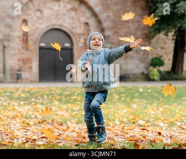Kind in warmer Kleidung genießt Herbstlaub. Spielerischer Outdoor-Moment, unbeschwerter Spaß, gesunde Kindheit Stockfoto
