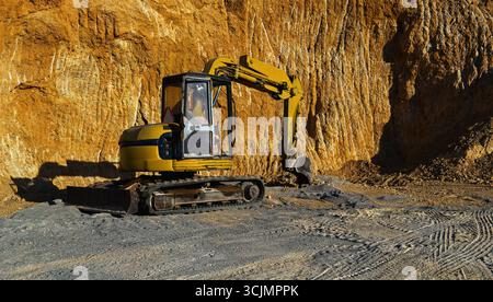 Bagger, der auf dem Steinbruchgelände mit felsigen Klippen und Reifenspuren auf dem Boden arbeitet Stockfoto