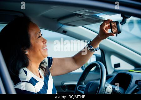 Lächelnde, selbstbewusste asiatische Frau mittleren Alters im Auto, die in den Rückspiegel blickt und Selbstvertrauen, Vielfalt und modernen urbanen Lebensstil symbolisiert. Stockfoto