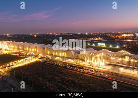 Aus der Vogelperspektive des Bahnhofs Stazione AV Mediopadana mit seinem gewellten Dach, das warm gegen den Abendhimmel leuchtet, Stazione AV Mediopadana, Emilia-Romagna, Italien. Stockfoto