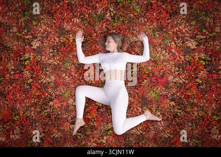 Eine Frau in einem weißen Yoga-Outfit spielt in einer beruhigenden Pose, während sie auf einem Bett aus bunten Herbstblättern liegt. Die Szene fängt die Schönheit der Natur ein Stockfoto