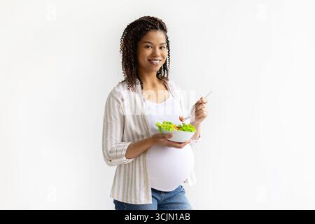 Lächelnde schwarze schwangere Dame, die eine Schüssel mit frischem Gemüsesalat und Gabel hält Stockfoto