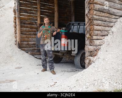 Porträt eines ukrainischen Soldaten in taktischer Ausrüstung, der seinen Helm hält und neben einer getarnten Ausgrabung mit einem Auto steht. Krieg in der Ukraine. Stockfoto