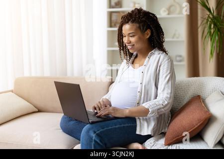 Glückliche schwangere schwarze Frau sitzt auf dem Sofa und arbeitet zu Hause am Laptop Stockfoto