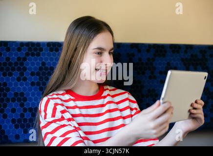 Ein Teenager mit gestreiftem Hemd lächelt, während sie im Zug etwas auf ihrem Tablet ansieht, was Spaß, digitale Verbindung und Entspannung zeigt Stockfoto