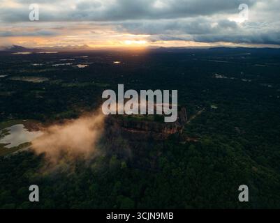 Blick aus der Vogelperspektive auf Sigiriya, eine majestätische Felsenfestung, die durch die grüne Landschaft unter einem dramatischen Sonnenuntergangshimmel durchsticht, Lion Rock, Zentralprovinz, Sri Lanka. Stockfoto