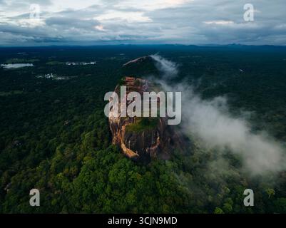 Blick aus der Vogelperspektive auf eine majestätische Felsenfestung, die durch eine Nebeldecke mitten im üppigen Grün durchsticht, Lion Rock, Zentralprovinz, Sri Lanka. Stockfoto
