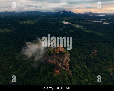 Blick aus der Vogelperspektive auf die Felsfestung Sigiriya, die durch das smaragdgrüne Baldachin durchsticht, gekrönt von antiken Ruinen unter einem brütenden Himmel, Lion Rock, Zentralprovinz, Sri Lanka. Stockfoto