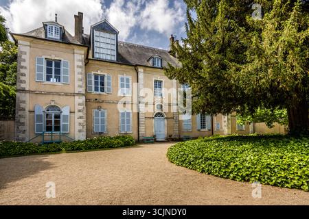 Das George-Sand-Haus (Maison de George Sand) in Nohant, Gemeinde Nohant-Vic, im Berry, Département Indre, Centre-Val de Loire, Frankreich, zeigt seine Eingangsfassade zum Kieshof. Das zweistöckige Herrenhaus hat hellblaue Fensterläden, Naturstein-Ecken und ein Schieferdach mit Gauben und Schornsteinen. Zwei grüne Bänke stehen neben der Haustür, und Beete mit niedrigen Sträuchern und Efeu säumen den Hof. Rechts erhebt sich ein großer Nadelbaum über die Fassade (möglicherweise die große Eibe, die den Haupthof beschattet). Das Herrenhaus stammt aus den 1770er Jahren und hat einen klassischen Grundriss im Louis Stockfoto