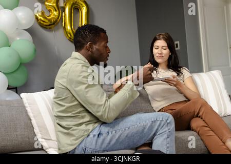 Afroamerikanischer Mann und afroamerikanische Frau genießen Kuchen auf der Couch während der Geburtstagsfeier Stockfoto